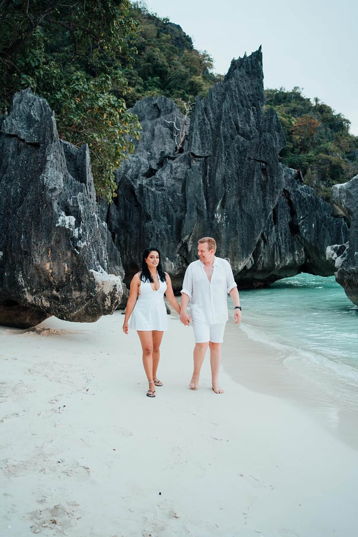 A couple in white outfits walks hand in hand along a sandy stretch of Banol Beach, with turquoise water gently lapping at their feet. Jagged rock formations and lush green foliage are in the background, creating a serene, romantic atmosphere perfect for daydreaming about island hopping adventures.
