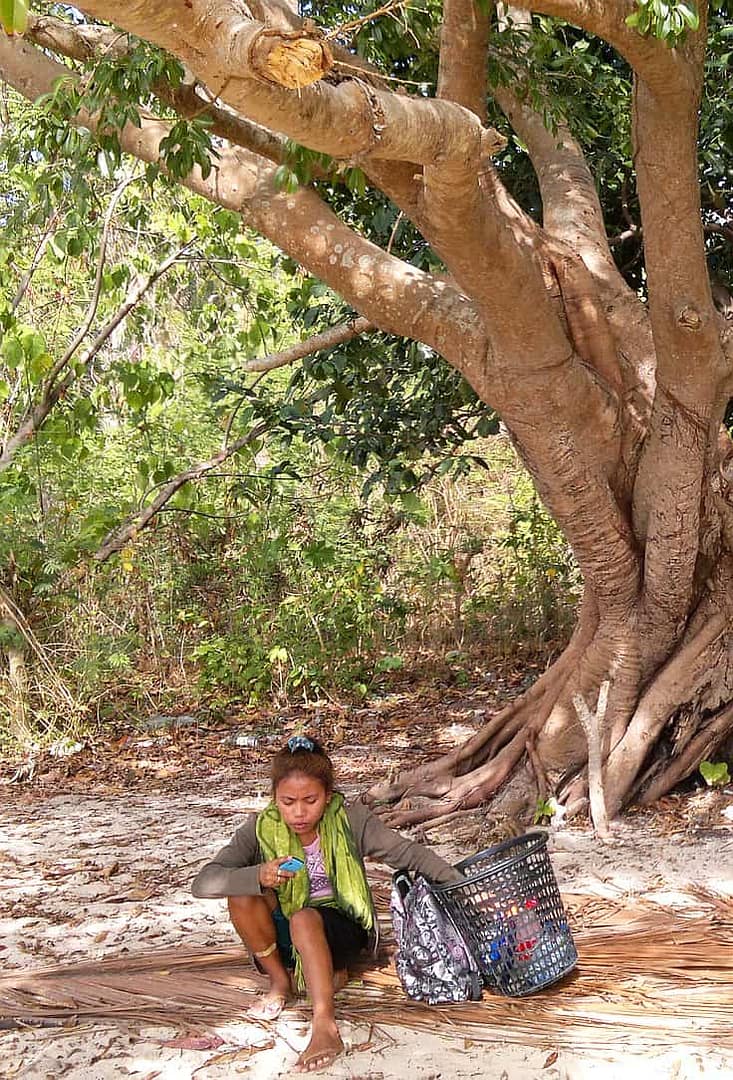 A person sits under a large, leafy tree in a sunny, forested area reminiscent of island hopping. They are holding a drink and have a green scarf around their neck. Beside them are two large baskets. The ground is covered with dry leaves.