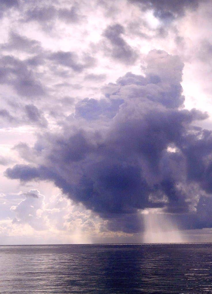 Dramatic cloud formations loom over a calm ocean at sunrise, with rays of light streaming through gaps in the clouds, creating a serene and majestic seascape perfect for island hopping or captivating boat tours.