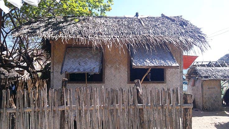 A small, thatched-roof house with woven bamboo walls and two open windows sits peacefully, echoing the charm of island hopping adventures. A wooden fence encloses the area while a tree provides shade on one side under a clear blue sky.