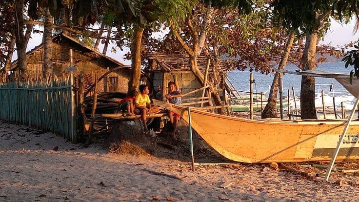 Two children sit on a log under trees near a small wooden house by the beach, perfect for starting island hopping adventures. A boat awaits nearby on the sand, and the sea shimmers in the warm glow of sunset, painting an idyllic scene.