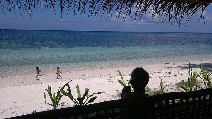 A person sits under a thatched roof, watching two children run on a sandy beach near the turquoise sea, dreaming of island hopping. Green plants frame the foreground, and the horizon is visible under a partly cloudy sky.