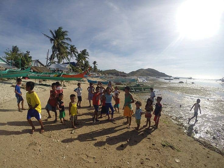 A group of children play on a sandy beach under the bright sun. Fishing boats hint at upcoming island hopping adventures, while palm trees sway in the background against a hilly landscape. The sea is at low tide.