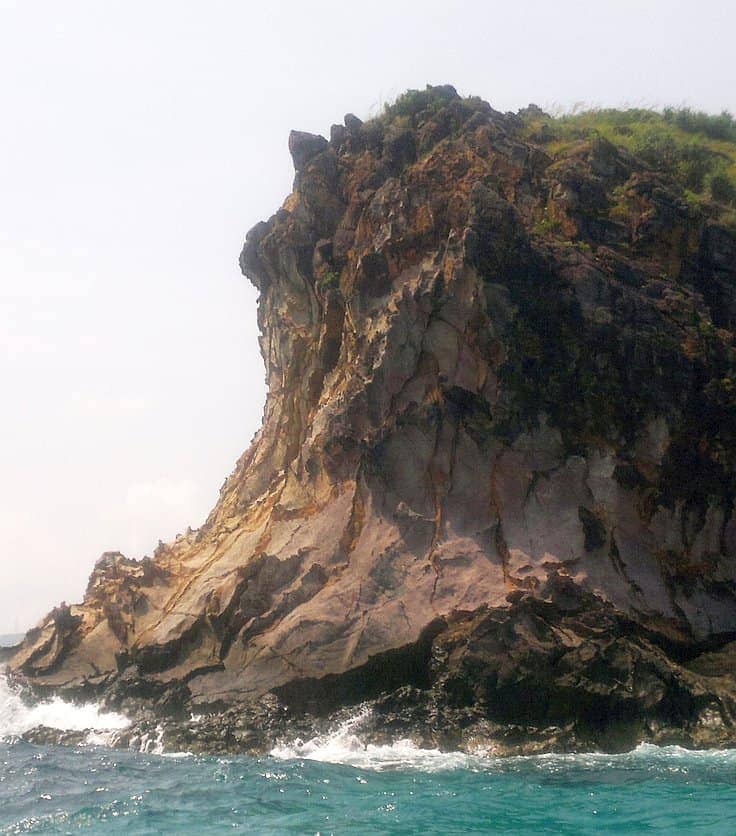 A rocky cliff juts out into the sea, waves crashing against its base as boat tours pass by. The cliff, topped with grass and plants, stands boldly under a cloudy sky and overlooks the blue ocean waters—a perfect stop on an island hopping adventure.