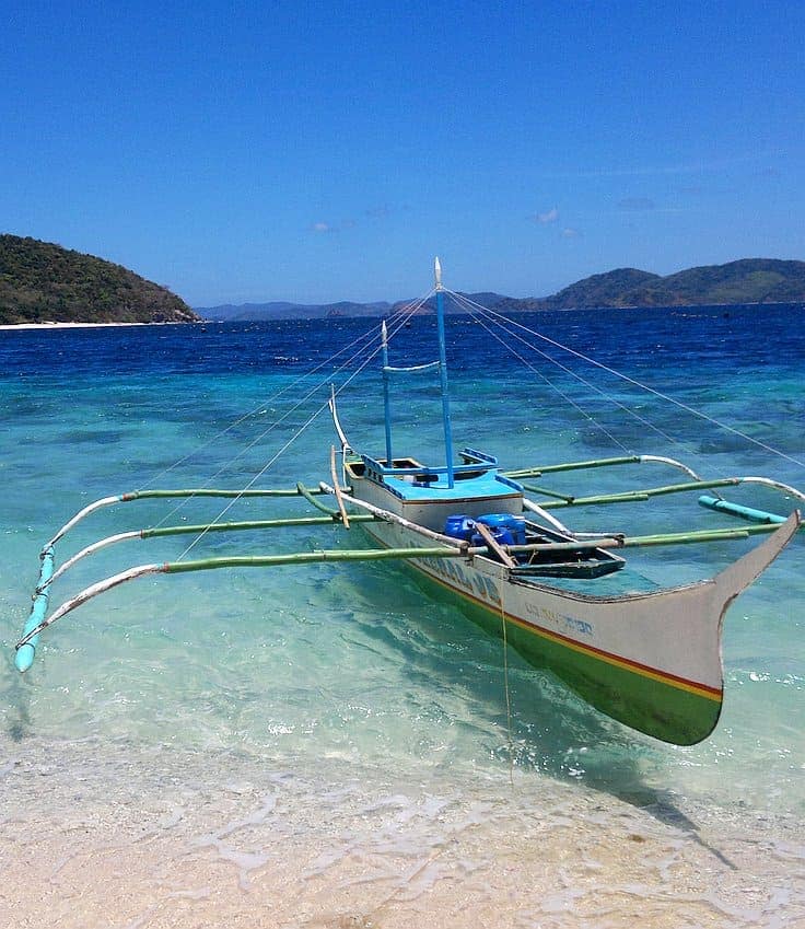 A traditional outrigger boat, perfect for island hopping adventures, rests on a pristine, turquoise shoreline under a clear blue sky, surrounded by distant green islands.