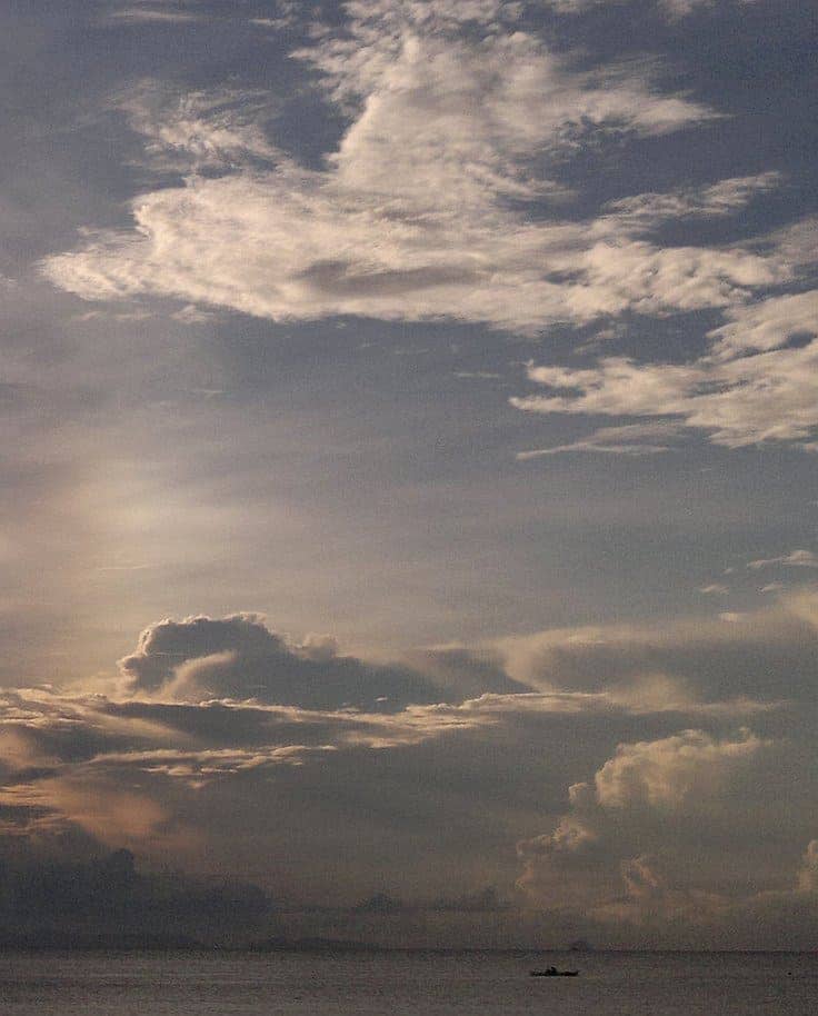 A small boat sails on the horizon under a vast sky filled with dramatic clouds at sunrise or sunset. The scene evokes tranquility, as soft light illuminates the clouds, reminiscent of serene island hopping adventures.