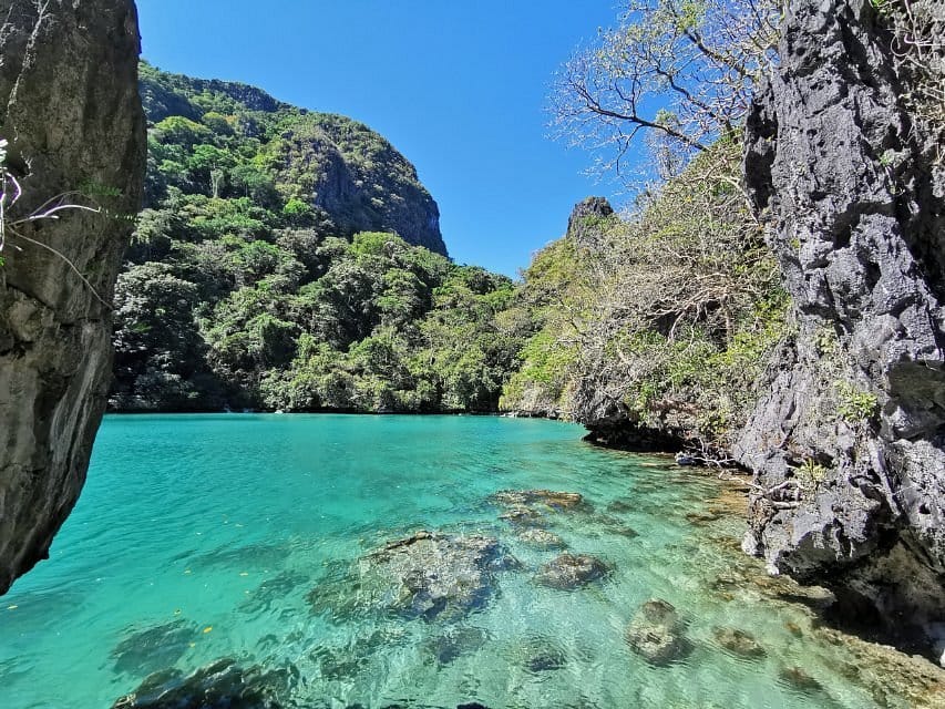 A tranquil turquoise lagoon, reminiscent of the Big Lagoon in Palawan, is surrounded by rocky cliffs and lush green vegetation under a clear blue sky. The transparent water reveals rocks beneath the surface, crafting a serene and picturesque natural scenery perfect for island hopping enthusiasts.