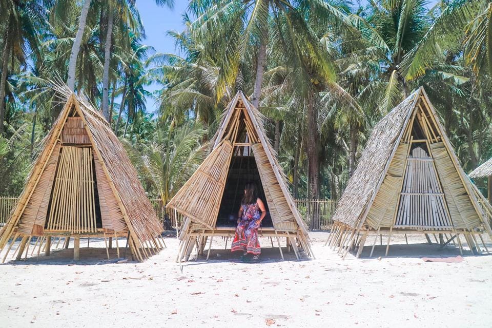 Three triangular bamboo huts stand on a sandy beach in Palawan, surrounded by palm trees under a clear blue sky. Inside the center hut, a person in colorful clothing is seated, partially shaded by the structure—planning a surprise proposal during their romantic vacation.