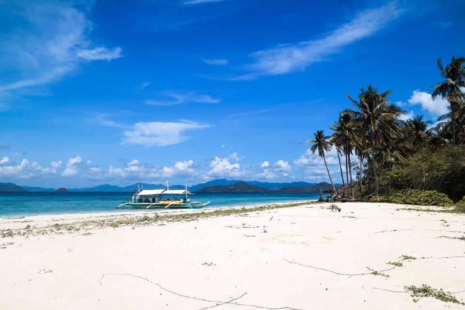 A secluded beach with white sand stretches out to a turquoise sea, perfect for a romantic vacation. A traditional boat is anchored, and the beach is lined with tall palm trees under a clear blue sky, with distant islands visible across the water.