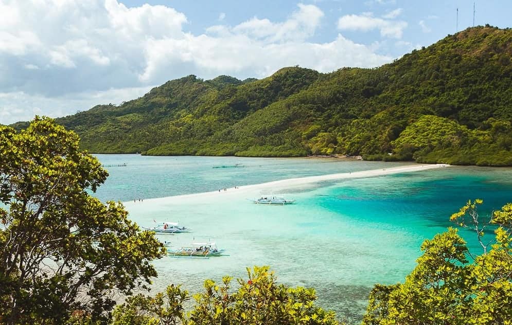 A scenic view of a tropical island with lush green hills and a narrow sandbar extending into the turquoise sea. Several small boats from a Palawan boat tour float near the sandbar under a partly cloudy sky, capturing the essence of paradise.