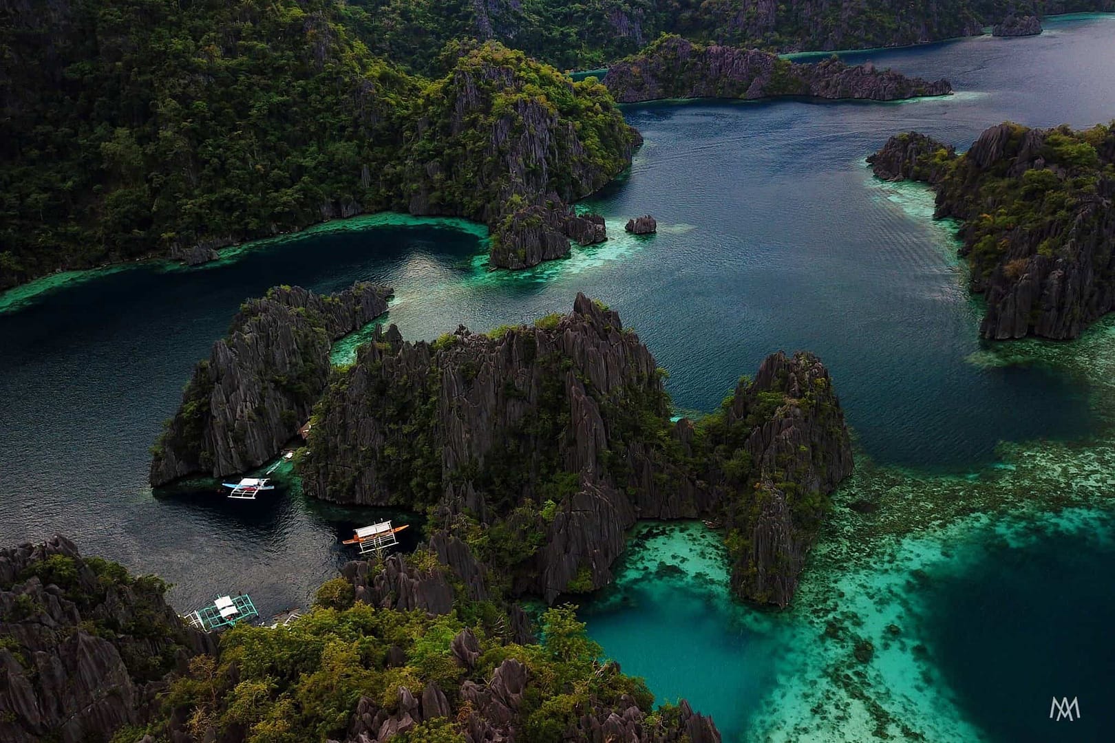 A stunning aerial view of cliffs and turquoise waters showcases boats anchored near rocky islets, perfect for island hopping in El Nido. Surrounded by lush greenery, the scene captures a vibrant contrast between the dark waters and vivid green landscapes.