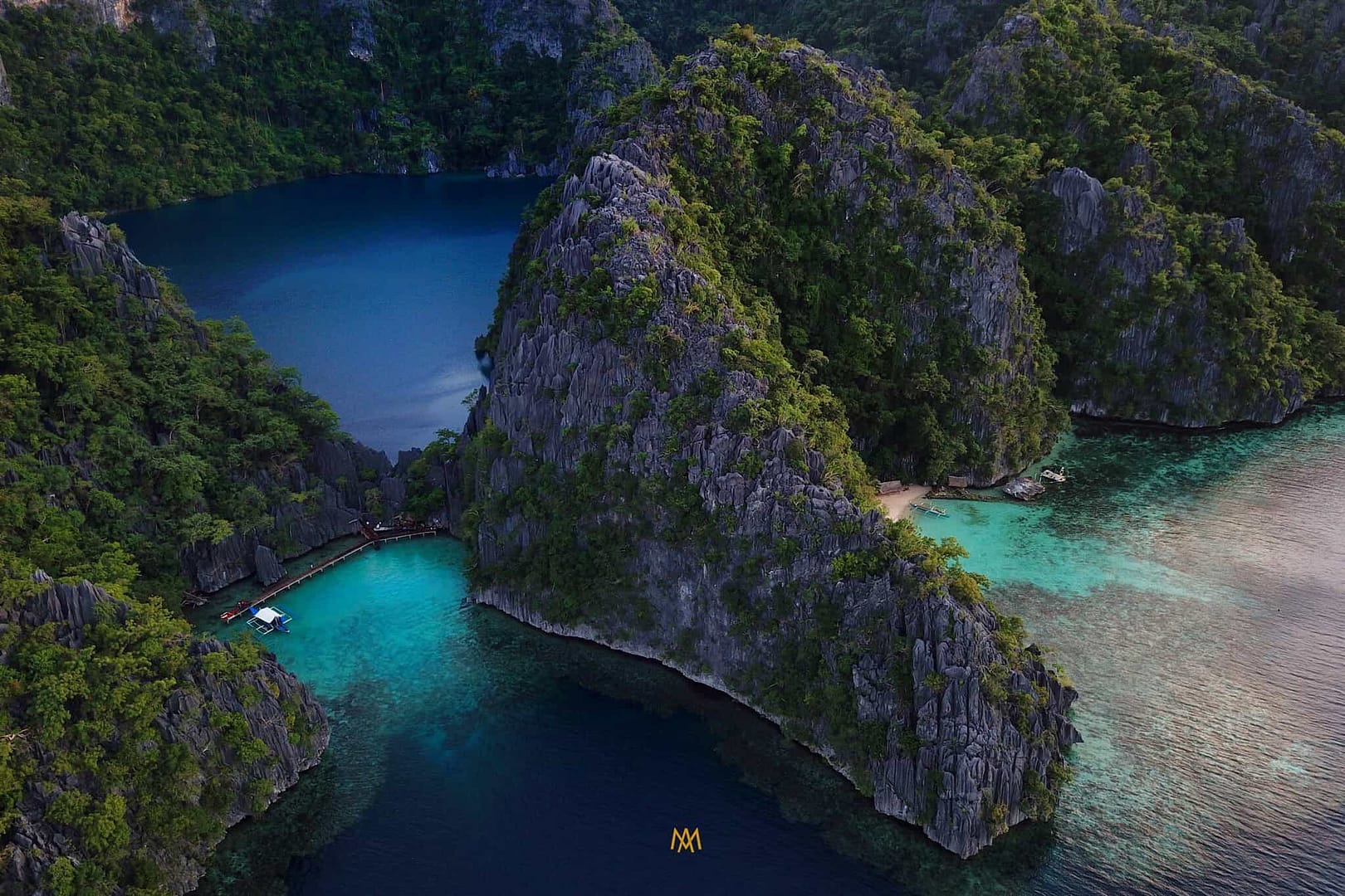 Aerial view of a tropical island in El Nido, with steep, rocky cliffs covered in lush greenery. The island is surrounded by vibrant blue and turquoise waters perfect for boat tours. A wooden dock extends over the water, connecting to a small boat ideal for island hopping adventures.